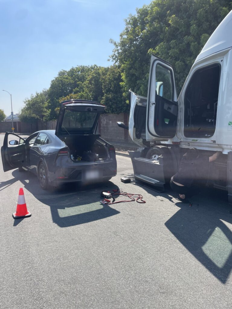 Tráiler blanco con las puertas de la cabina abiertas y un técnico agachado junto a la cabina; en el pavimento hay cables para pasar corriente y un arrancador portátil, junto a un Prius con la cajuela abierta y un cono
