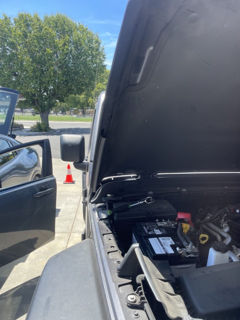 Jeep Wrangler seen over the open hood, battery with a red terminal cover under the raised hood, a traffic cone and a second car in the background